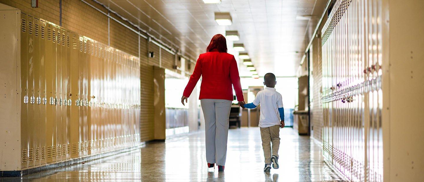 An adult and a child holding hands, walking down a hallway full of lockers. 