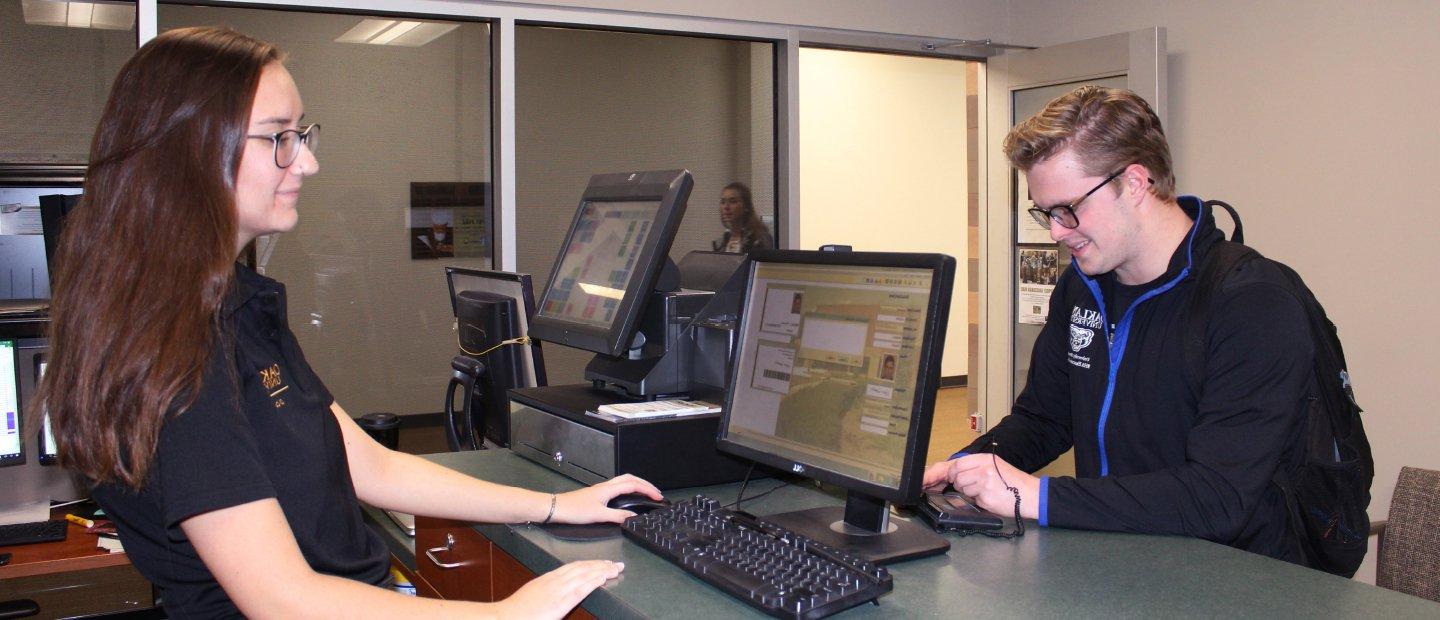 young woman standing behind a desk with a computer, assisting a young man on the other side of the counter